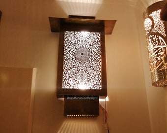 May include: A brass wall sconce with intricate cut-out designs, casting a warm glow on the wall. The sconce is illuminated from within, creating a beautiful pattern of light and shadow. The text "ShopMoroccanDesigns" is visible on the sconce.