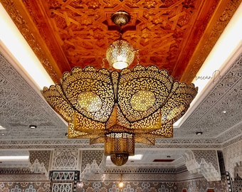 May include: A large, ornate, gold-colored chandelier with intricate, lacy designs hangs from a ceiling with a carved wooden pattern. The chandelier has multiple lights and is illuminated, casting a warm glow.