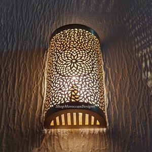 Könnte beinhalten: Eine goldene Wandleuchte aus Metall mit einem dekorativen, filigranen Ausschnittmuster. Die Leuchte ist beleuchtet und strahlt ein warmes Licht ab. Der Text "ShopMoroccanDesigns" ist auf der Leuchte sichtbar.