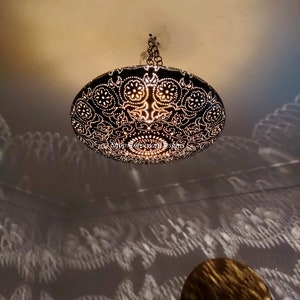 May include: A black metal ceiling light fixture with a decorative, intricate pattern. The light is on and casting a shadow on the ceiling.