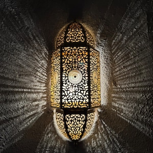 May include: Ornate, gold-colored metal lantern with intricate cut-out patterns, casting decorative light and shadow patterns on a textured wall. The lantern has a central light source and a detailed, geometric design.