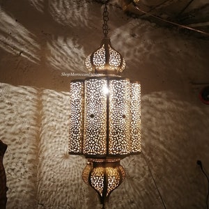 May include: A gold Moroccan pendant light with intricate cut-out designs. The light is hanging from a chain and has a round top and a cylindrical body.