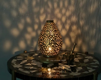 May include: A gold metal table lamp with a decorative, intricate, cut-out design. The lamp is on a round, black and white tiled table. The text "Shop Moroccan Designs" is visible on the table.