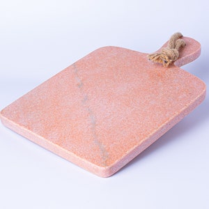 May include: A pink marble chopping board with a rope handle. The board is textured and has a light pink colour with white streaks.