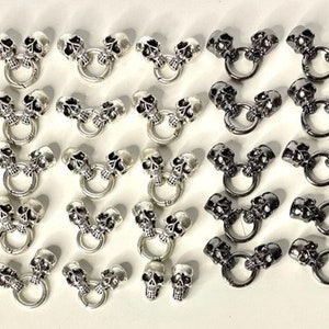 May include: A collection of silver and black skull charm pendants with a ring attachment. The charms are in various designs, some with two skulls and others with a single skull.