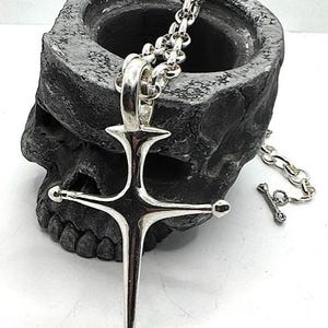 May include: A silver-colored pendant necklace featuring a stylized cross design. The cross has rounded tips and is suspended from a chain. The necklace is displayed against a dark, textured background that resembles a skull.