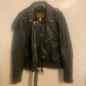 以下が含まれることがあります： 黒い革のオートバイジャケット。ジッパー開閉、複数のポケット、ベルト付き。ノッチカラーと肩のエポレットを備えたクラシックなバイカースタイル。ラベルには「Protec Leather Apparel」と記載されています。