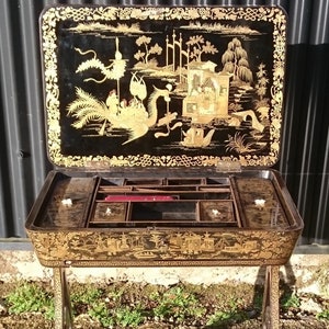 May include: Antique black and gold lacquered writing table with a hinged top and multiple drawers. The top features a detailed Asian-inspired scene with gold accents.