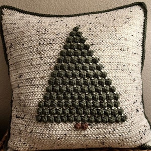 May include: A white and green crocheted pillow with a green Christmas tree design. The tree is made of textured stitches and has a brown felt trunk.