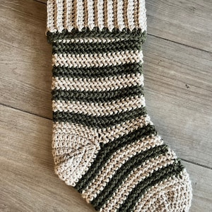 Könnte beinhalten: Ein gehäkelter Weihnachtssocken mit einem grün-beigen Streifenmuster. Der Socken hat einen beigefarbenen Bund.
