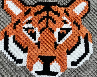 May include: A crocheted tiger face with orange, white, and black yarn. The tiger is on a gray background.