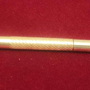 May include: A cylindrical gold-colored metal tool featuring a central section with a textured, diagonal ribbed pattern and smooth, polished ends. The object is straight and uniform in shape, displaying a metallic finish throughout its length.