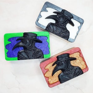 May include: Three rectangular soap bars featuring a black plague doctor design. The soaps are displayed on a white surface. The soaps have different coloured backgrounds: green, white, and orange.