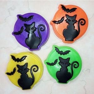 May include: Four round, colorful soap bars with a black cat and bats design. The soaps are purple, orange, green, and yellow.