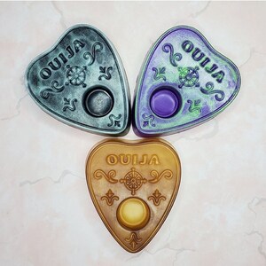 May include: Three heart-shaped, tealight-style candle holders with the word "OUIJA" written on them. The candle holders are in shades of green, purple, and black.