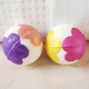 Peut inclure: Deux bombes de bain avec une base blanche et des motifs floraux colorés. Une bombe de bain a des fleurs violettes, orange et dorées. L'autre bombe de bain a des fleurs roses et jaunes.