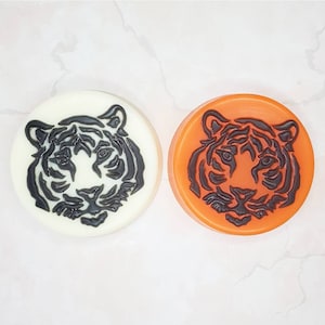May include: Two round soap bars with a black tiger face design. One soap bar is white and the other is orange.