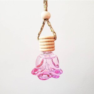 May include: A pink, flower-shaped glass diffuser bottle with a wooden cap and a braided rope hanger. The bottle is filled with a pink liquid. The wooden cap has a series of wooden rings. A wooden bead is at the top of the rope.
