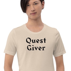 May include: A beige short-sleeved t-shirt with the text "Quest Giver" printed in black.