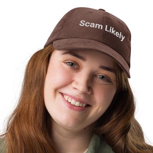May include: A brown corduroy baseball cap with the text "Scam Likely" embroidered in white. The cap is worn by a person wearing a green corduroy jacket.