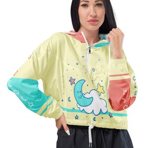 May include: A cropped, yellow windbreaker jacket with a whimsical design. The jacket features a smiling moon and cloud graphic, along with stars. It has teal and coral accents on the sleeves and hood, and a drawstring waist.