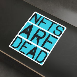 May include: A black and white sticker on a skateboard with a blue background. The sticker says "NFTs ARE DEAD" in a bold, blocky font.