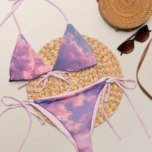 Puede incluir: Un bikini rosa y morado con estampado de nubes. La parte superior del bikini es de estilo triangular y la parte inferior es de estilo de amarre lateral. El bikini está sobre una estera de paja tejida.
