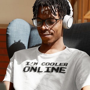 Può includere: Una persona indossa una maglietta bianca con il testo "I'M COOLER ONLINE" in lettere pixelate nere. La persona è seduta su una sedia e indossa delle cuffie, guardando un laptop.