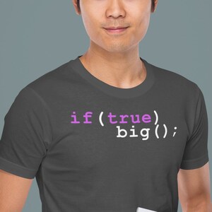Może przedstawiać: Mężczyzna w ciemnoszarym t-shircie z nadrukiem kodu "if (true) big();" w kolorze białym z różowymi akcentami. Trzyma srebrny laptop w lewej ręce.