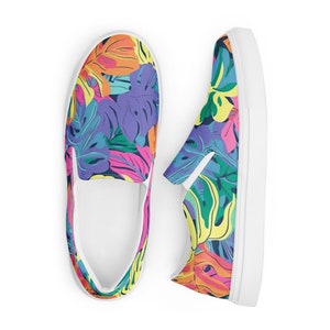 K&ouml;nnte beinhalten: Ein Paar wei&szlig;e Slip-on-Canvas-Schuhe mit einem bunten tropischen Blatt-Muster. Die Schuhe haben eine wei&szlig;e Sohle und ein wei&szlig;es Gummiband oben.