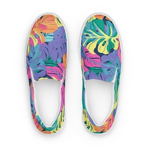 K&ouml;nnte beinhalten: Ein Paar wei&szlig;e Slip-on-Canvas-Schuhe mit einem bunten tropischen Bl&auml;ttermuster. Das Muster zeigt leuchtend rosa, gelbe, gr&uuml;ne, blaue und orange Bl&auml;tter.