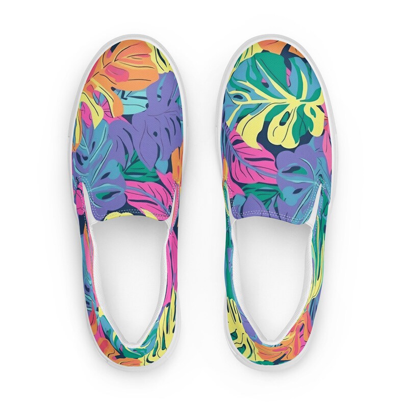 K&ouml;nnte beinhalten: Ein Paar wei&szlig;e Slip-on-Canvas-Schuhe mit einem bunten tropischen Bl&auml;ttermuster. Das Muster zeigt leuchtend rosa, gelbe, gr&uuml;ne, blaue und orange Bl&auml;tter.