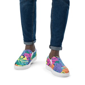 K&ouml;nnte beinhalten: Ein Paar wei&szlig;e Canvas-Slipper mit einem bunten tropischen Bl&auml;tterdruck. Die Schuhe haben eine wei&szlig;e Sohle und werden von einer Person in blauer Jeans getragen.