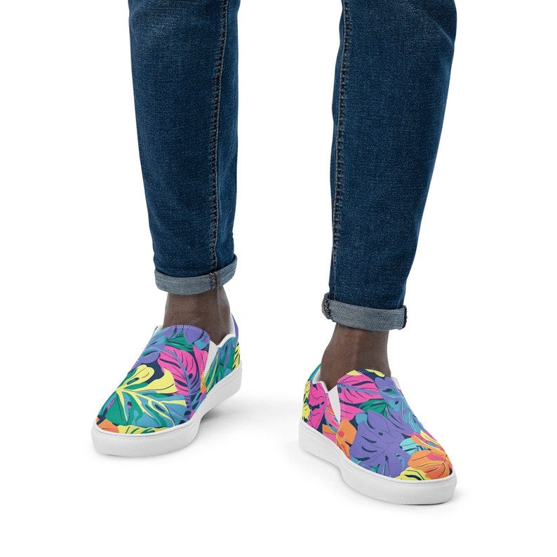 K&ouml;nnte beinhalten: Ein Paar wei&szlig;e Canvas-Slipper mit einem bunten tropischen Bl&auml;tterdruck. Die Schuhe haben eine wei&szlig;e Sohle und werden von einer Person in blauer Jeans getragen.