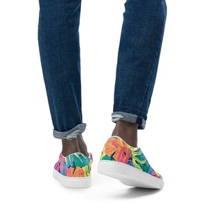 K&ouml;nnte beinhalten: Ein Paar wei&szlig;e Canvas-Sneaker mit niedrigem Schaft und einem bunten tropischen Blattdruck. Die Sneaker haben eine wei&szlig;e Gummisohle und werden von einer Person getragen, die blaue Jeans tr&auml;gt.