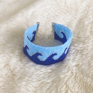 May include: A beaded bracelet featuring a wave pattern in shades of blue. The bracelet has a light blue background with dark blue waves. The clasp is silver-toned, and the bracelet is displayed on a fluffy, white surface.