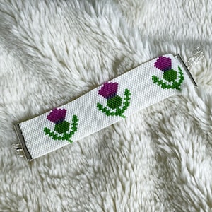 May include: A white beaded bracelet featuring a repeating pattern of purple and green thistle flowers. The bracelet has a silver-colored clasp and is displayed on a white, fluffy surface. The bracelet is approximately 1 inch wide.