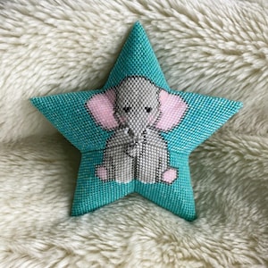 Op de afbeelding: Een tealkleurig, stervormig sierkussen met een kruissteekontwerp van een olifant. De olifant is grijs met roze oren en voeten. Het kussen is ongeveer 20 cm breed en ligt op een zacht, wit oppervlak.