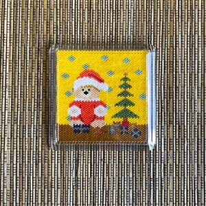May include: A square coaster featuring a cross-stitch design of a teddy bear wearing a red and white Santa hat and outfit, standing next to a green Christmas tree with small gifts on a yellow background with blue snowflakes.