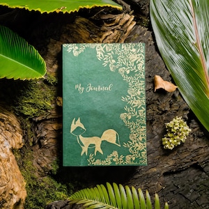 May include: A rectangular green journal with a gold foil illustration of a fox and floral vines along the right edge. The text My Journal is printed in gold script on the front cover. The cover features a matte texture and a solid green colour.