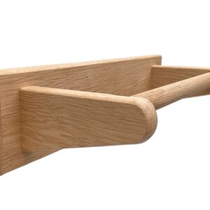 Puede incluir: Un porta rollo de papel de pared de madera con un acabado natural. El soporte tiene una base rectangular y una barra redondeada para sujetar el rollo.
