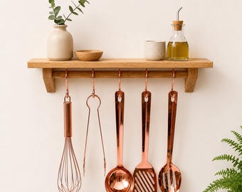 Puede incluir: Una estantería de madera con utensilios de cocina de color cobre colgando debajo. Los utensilios incluyen un batidor, unas pinzas, un cucharón, una espátula ranurada y una cuchara. Un pequeño jarrón con vegetación, un cuenco de madera, una taza de cerámica y una botella de vidrio con aceite están en el estante.