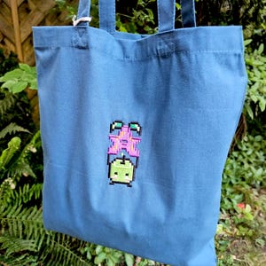 May include: A blue tote bag featuring an embroidered pixel art design of a green character with a pink crown. The bag has two handles and is made of a durable fabric. The design is on the front.