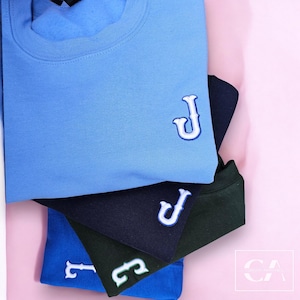 Könnte beinhalten: Ein Stapel von vier T-Shirts in verschiedenen Farben, jedes mit einem weißen gestickten Buchstaben "J" auf der Vorderseite. Die Shirts sind hellblau, dunkelblau, grün und royalblau.