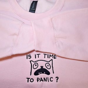 May include: A light pink sweatshirt with the text "IS IT TIME TO PANIC?" and a cartoon cat face with wide eyes and an open mouth. The sweatshirt is made of a soft material and has long sleeves.