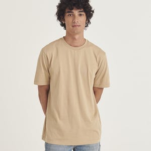 Op de afbeelding: Een effen beige T-shirt met ronde hals. Het shirt met korte mouwen is effen van kleur en wordt gedragen met een blauwe spijkerbroek. Het model staat voor een witte achtergrond.