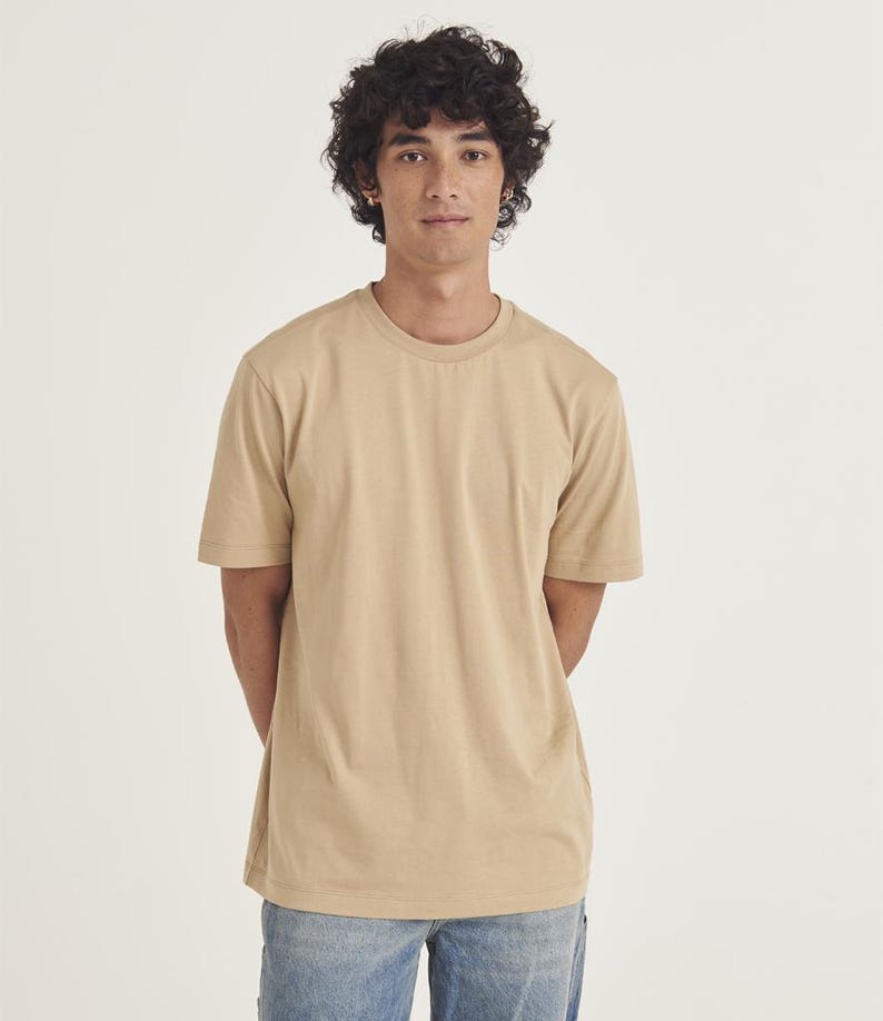 Op de afbeelding: Een effen beige T-shirt met ronde hals. Het shirt met korte mouwen is effen van kleur en wordt gedragen met een blauwe spijkerbroek. Het model staat voor een witte achtergrond.