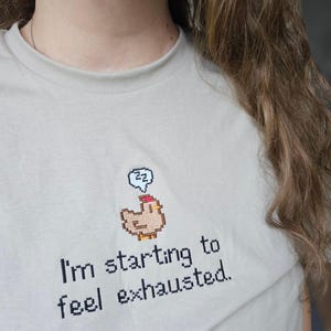 Op de afbeelding: Lichtbeige T-shirt met de geborduurde tekst "I'm starting to feel exhausted." Boven de tekst staat een gepixeleerde kip met een slaapbel. Het shirt heeft een ronde hals en korte mouwen.
