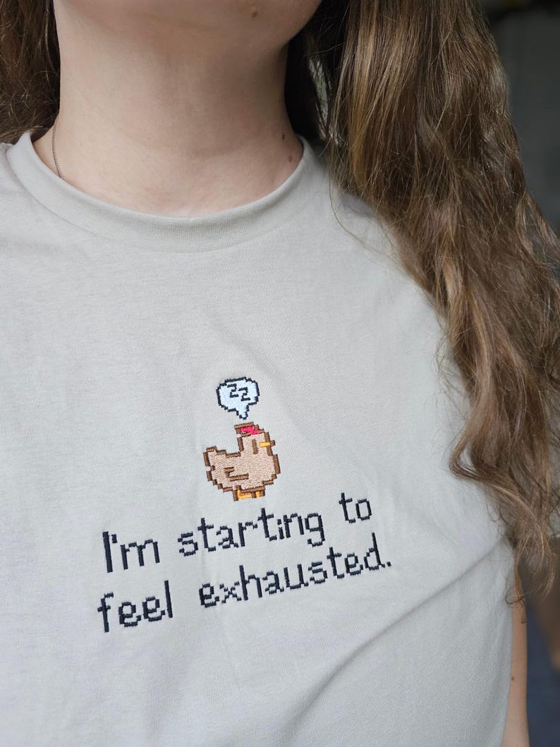 Op de afbeelding: Lichtbeige T-shirt met de geborduurde tekst "I'm starting to feel exhausted." Boven de tekst staat een gepixeleerde kip met een slaapbel. Het shirt heeft een ronde hals en korte mouwen.