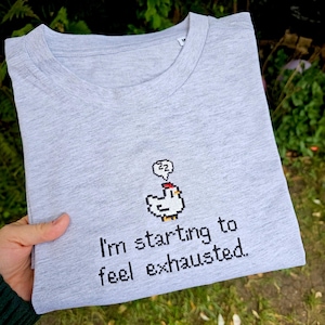 Puede incluir: Camiseta gris jaspeado con un gráfico de pollo pixelado y el texto "I'm starting to feel exhausted". El pollo tiene una cresta roja y una burbuja de pensamiento con "zz". La camiseta está doblada.